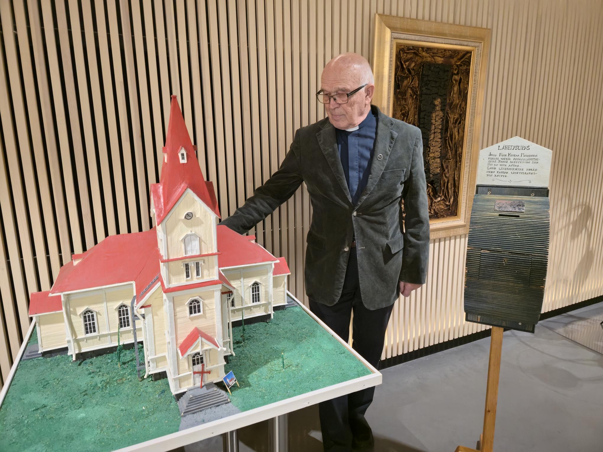 Esko Jaatinen, 90 vuotias syntymäpäiväsankari seisoo kirkon eteisessä vanhan kirkon pienoismallin ja malmbe...