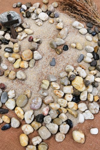 Kuvassa puinen ruskea risti, kiviä, hiekkaa ja heiniä