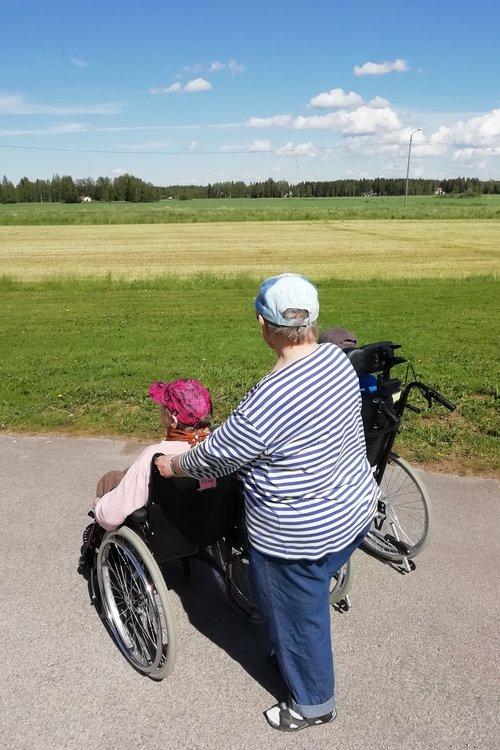Pyörätuoli ja avustaja kesäpäivänä katsomassa peltomaisemaa, pilviä taivaalla