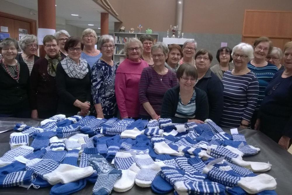 kymmeniä sinivalkoisia villasukkia pöydällä, jonka takana ryhmä sukat kutoneita naisia seisomassa